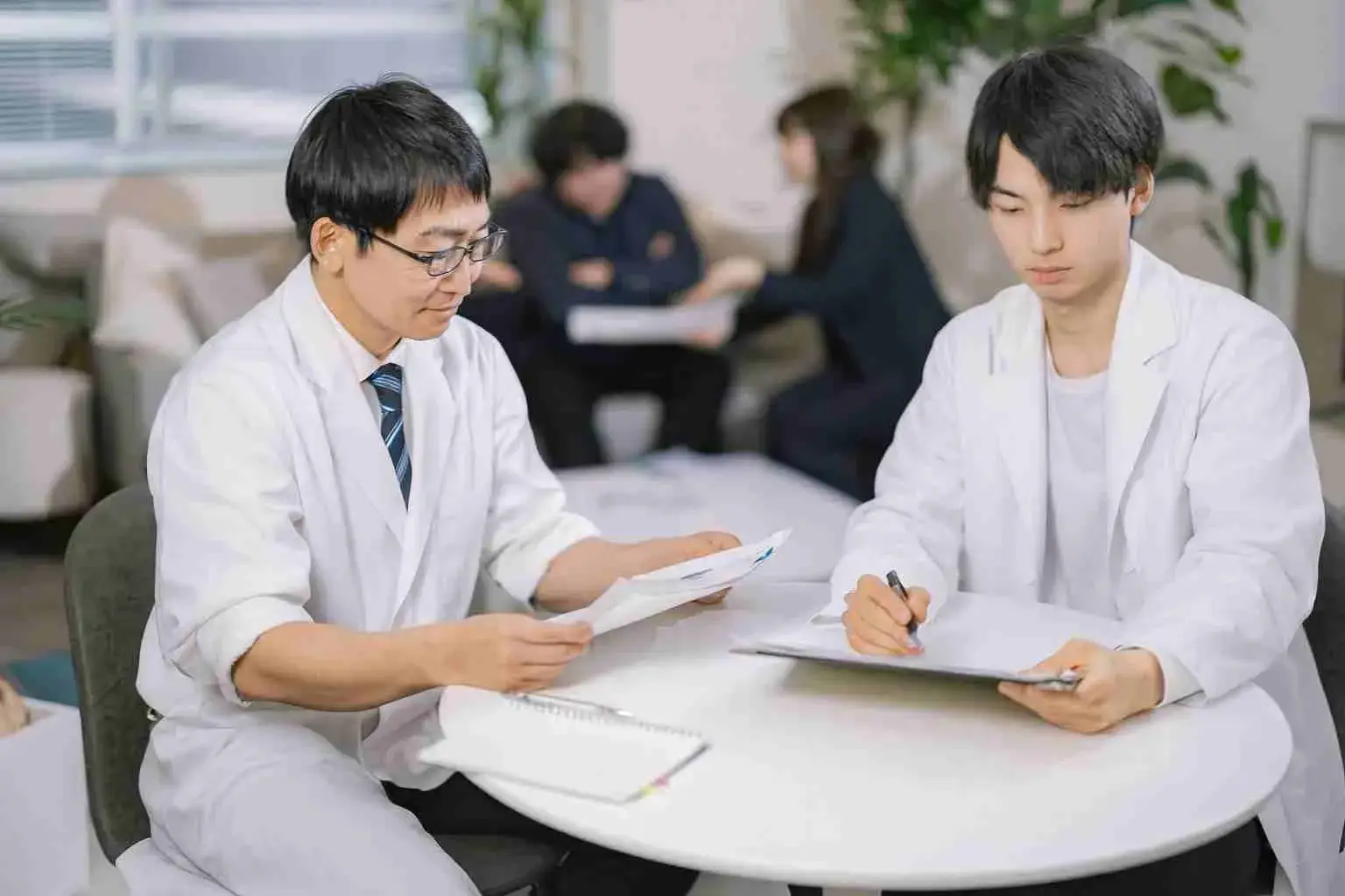 医療機関のミーティングで書類を確認する医師とスタッフ。背景には談笑する他のチームメンバーも映っている。