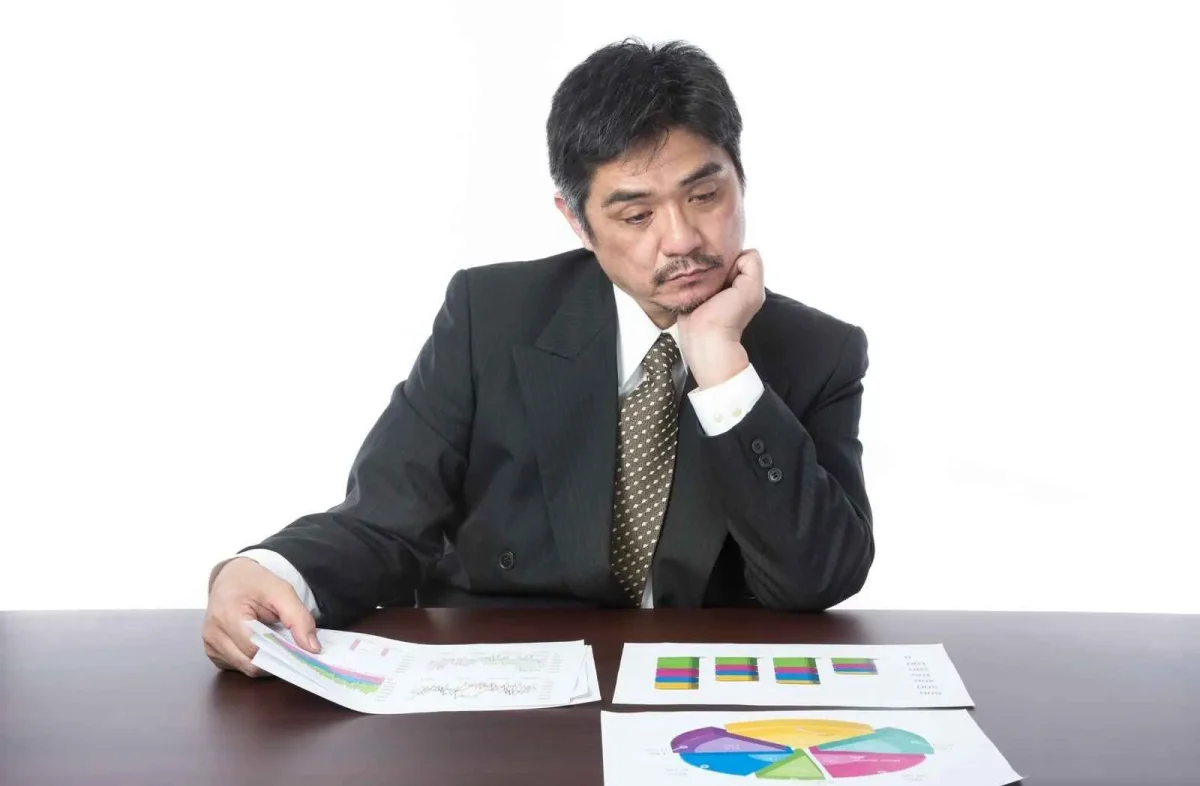 売上グラフや円グラフの資料を前に、頬杖をついて悩む経営者。経営分析や課題解決に取り組む様子。