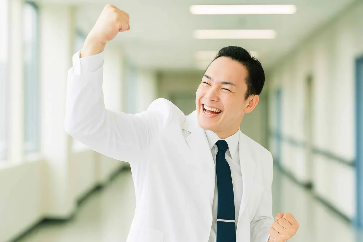 成功や目標達成を喜ぶ、笑顔でガッツポーズをする若い医師。明るい病院の廊下でのワンシーン。